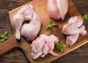 Assorted fresh raw chicken cuts including drumstick, breast, and wings arranged on a wooden cutting board with herbs