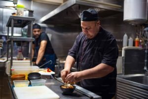 Professional chef plating a fresh dish in a busy commercial restaurant kitchen while another chef works in the background.