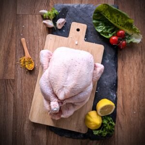 Whole fresh raw chicken on wooden chopping board with garlic, herbs, lemon, lettuce, and cherry tomatoes – premium poultry for restaurants and catering.