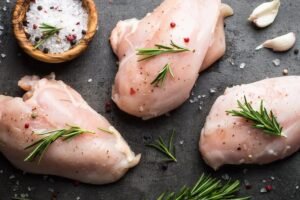 Raw chicken breasts arranged on a dark surface, seasoned with coarse salt, peppercorns, garlic cloves, and sprigs of fresh rosemary.