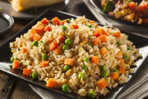 A plate of vegetable fried rice with diced carrots, green peas, and scrambled eggs, served on a dark square plate with a blurred background of side dishes.