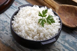 A bowl of fluffy white rice garnished with a parsley leaf, placed on a rustic wooden table with a wooden spoon beside it.