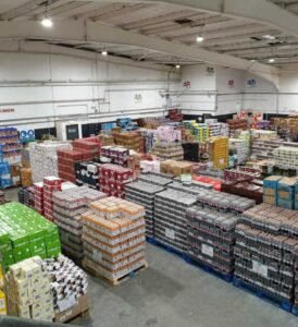 Spacious wholesale warehouse stocked with pallets of beverages, packaged foods, and bulk grocery products arranged in organized rows under bright industrial lighting.