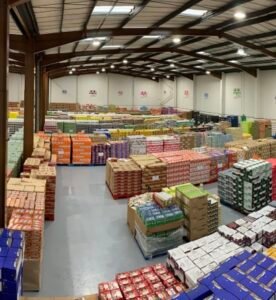 Large wholesale warehouse interior filled with stacked pallets and boxes of food and beverage products organized in rows under industrial lighting and high metal ceilings.