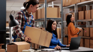 Warehouse employees organizing and handling cardboard boxes while using a laptop in a busy storage facility with shelves full of packaged goods and inventory.