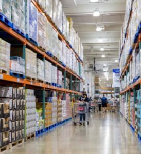 Long warehouse aisle lined with tall storage racks filled with bulk products and pallets, with shoppers and carts moving through the brightly lit wholesale distribution center.