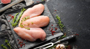 Three raw chicken breast fillets arranged on a black slate board with fresh herbs, peppercorns, and seasoning.